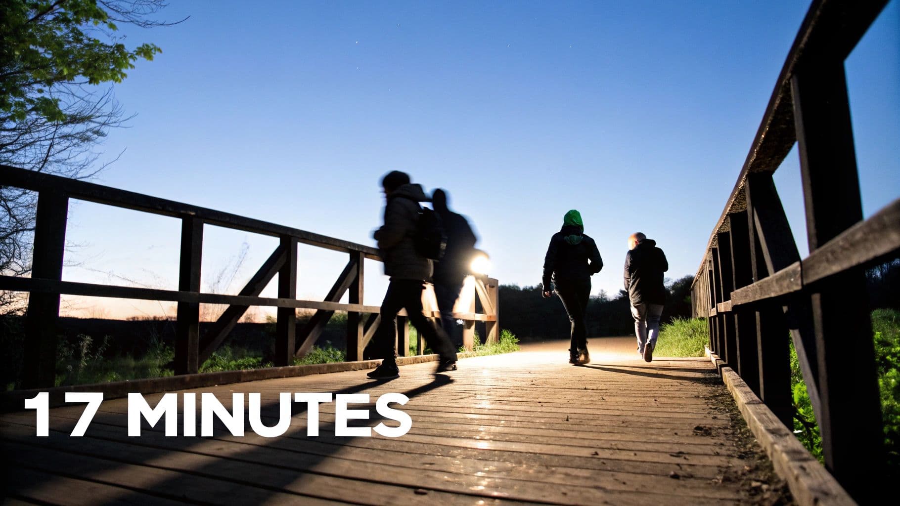Silhouettes of people walking on a wooden bridge at night under a blue sky, with a bright light.